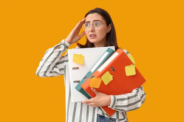 Stressed young businesswoman with document folders and sticky notes on yellow background