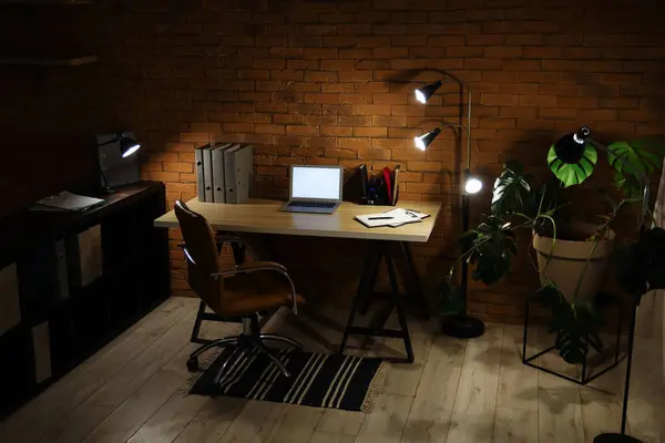 Interior of dark office with workplace, blank laptop and glowing lamps at night