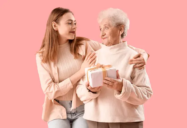 Young woman hugging her grandmother with gift on pink background