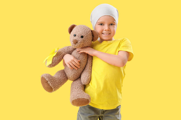 Cute little girl after chemotherapy with yellow ribbon and teddy bear on color background. Childhood cancer awareness concept