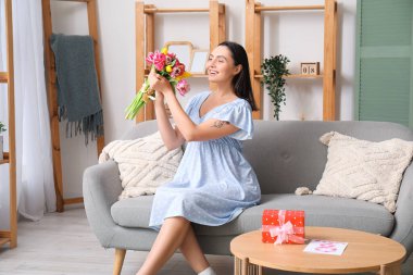 Evdeki kanepede güzel lalelerle dolu buketi olan mutlu genç bir kadın. Uluslararası Kadınlar Günü