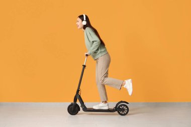 Kulaklıklı güzel bir Asyalı kadın sarı duvarın yanında modern elektrikli scooter kullanıyor.