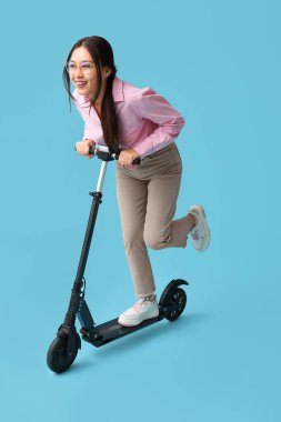 Güzel genç Asyalı kadın mavi arka planda modern elektrikli scooter kullanıyor.