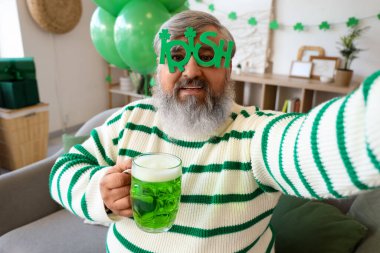 Aziz Patrick Günü 'nde gözlüklü ve biralı olgun bir adam evde selfie çekiyor.