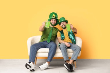 Yeşil sakallı, leprikon şapkalı mutlu genç adamlar ellerinde bira bardakları ve sarı duvarın yanındaki kanepede oturuyorlar. Aziz Patrick Günü kutlaması