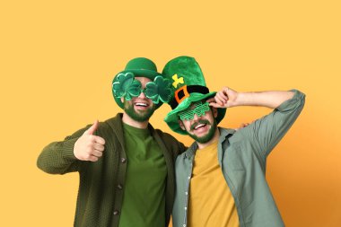 Leprikon şapkalı ve yeşil sakallı yonca şeklinde dekoratif gözlüklü genç adamlar sarı arka planda baş parmak hareketi yapıyorlar. Aziz Patrick Günü kutlaması