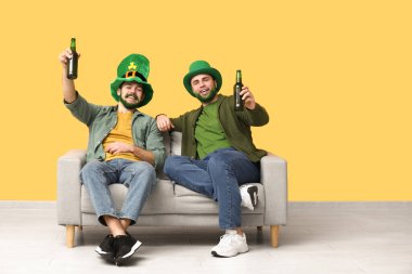 Yeşil sakallı ve yeşil sakallı mutlu genç adamlar ellerinde bira şişeleri ve sarı duvarın yanındaki kanepede oturuyorlar. Aziz Patrick Günü kutlaması