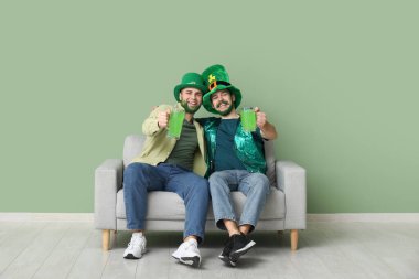 Yeşil sakallı, leprikon şapkalı genç adamlar ellerinde bira bardakları ve yeşil duvarın yanındaki kanepede oturuyorlar. Aziz Patrick Günü kutlaması