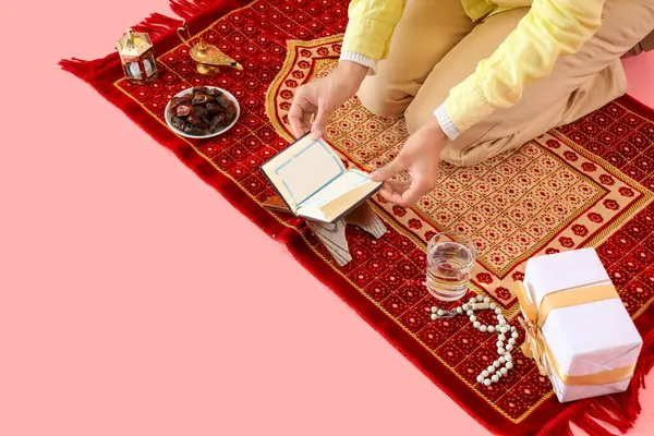 Muslim woman sitting on prayer mat with Ramadan symbols and Koran on pink background