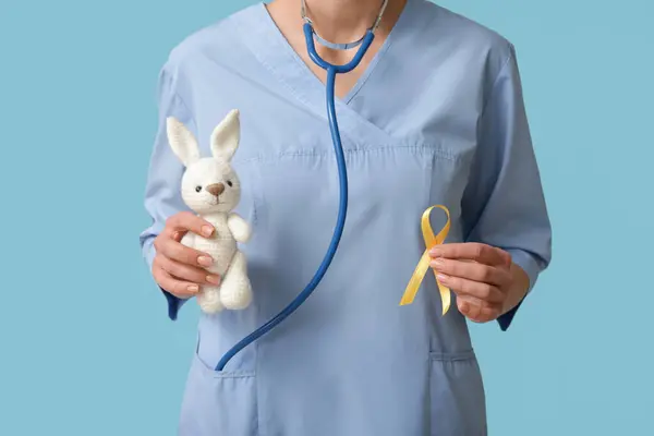 Doctor holding golden ribbon and toy bunny on blue background. Childhood cancer awareness concept