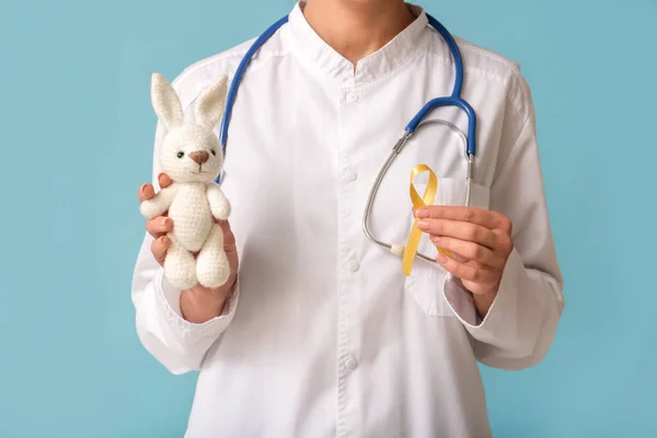Doctor holding golden ribbon and toy bunny on blue background. Childhood cancer awareness concept