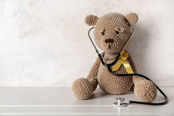 Toy bear with golden ribbon and stethoscope on white wooden table against grunge background. Childhood cancer awareness concept