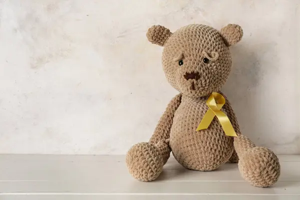 Toy bear with golden ribbon on white wooden table against grunge background. Childhood cancer awareness concept