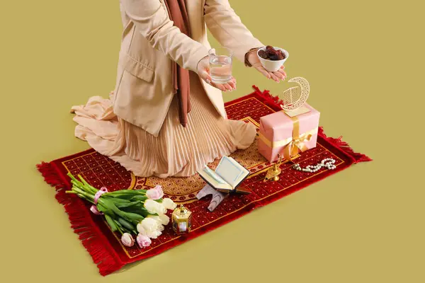 Mature Muslim woman with dates and glass of water on mat against green background. Ramadan celebration
