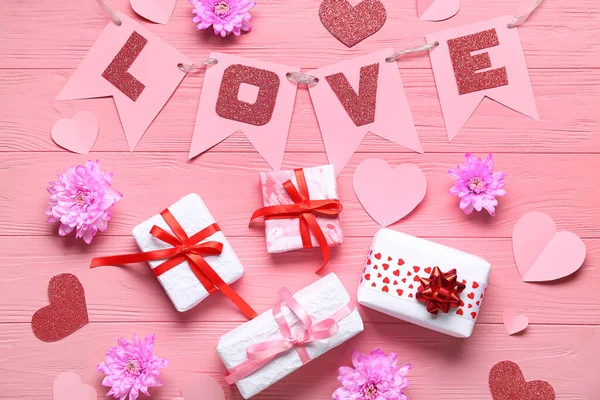 Garland made of paper flags with word LOVE, gift boxes and chrysanthemum flowers on pink wooden background. Valentine\'s Day celebration