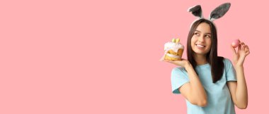 Tavşan kulaklı güzel bir genç kadın, pembe arka planda paskalya yumurtası ve pasta, mesaj için yer var.