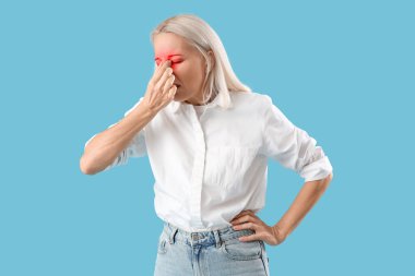 Açık mavi arka planda burnu ağrıyan olgun bir kadın.