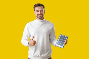 Portatif güneş paneli olan yakışıklı bir adam sarı arka planda başparmağını kaldırıyor.