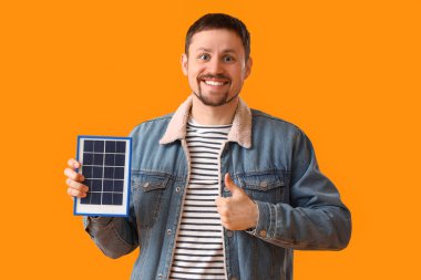 Portatif güneş paneli olan yakışıklı bir adam sarı arka planda başparmağını kaldırıyor.