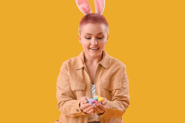 Young woman with bunny ears and Easter eggs on yellow background