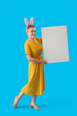 Tavşan kulaklı ve mavi arka planda boş posteri olan genç bir kadın. Paskalya kutlaması