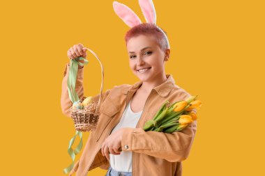 Tavşan kulaklı genç bir kadın, sarı arka planda bir sepet Paskalya yumurtası ve lalesi.
