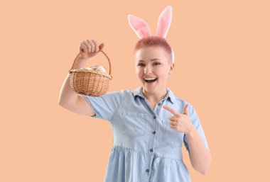 Tavşan kulaklı genç bir kadın bej arka planda paskalya yumurtası sepetine bakıyor.