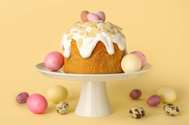 Stand with Easter cake and painted eggs on yellow background