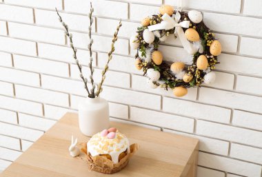 Vazoda söğüt dalları, beyaz tuğla duvarda Paskalya pastası ve çelenk olan bir masa.