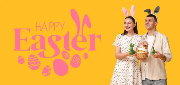 一对快乐的年轻夫妇 有小兔子耳朵 玩具和黄色背景的复活节篮子 复活节贺卡横幅 — 图库照片
