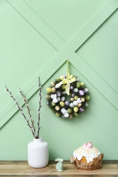 Table with willow branches in vase, Easter cake and wreath on green background