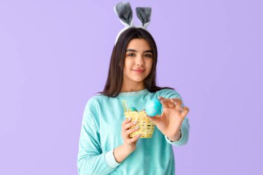 Leylak arka planında Paskalya yumurtası sepeti olan tavşan kulaklı genç bir kadın.