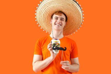 Sombrero giymiş Meksikalı genç bir adamın portresi, portakal rengi arka planda boyalı kafatası ve kağıt bıyıklı. Ölüler Günü Kutlaması