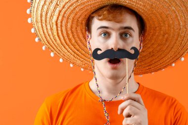 Sombrero giymiş, turuncu arka planda kağıt bıyıklı, şaşırmış Meksikalı bir adamın portresi.