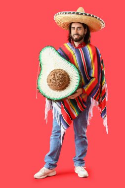 Kırmızı arka planda avokado şeklinde pinyatası olan yakışıklı Meksikalı genç adam.