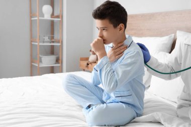 Doktor yatak odasında steteskobu olan hasta bir çocuğun ciğerlerini dinliyor.