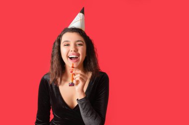 Kırmızı arka planda parti düdüğü çalan güzel genç bir kadın doğum gününü kutluyor.