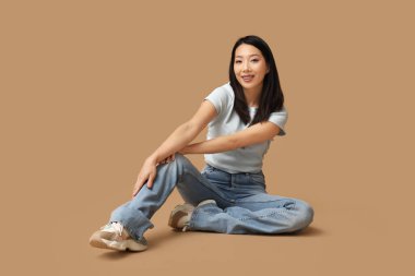 Kahverengi arka planda oturan şık kotlu güzel, mutlu Asyalı kadın.