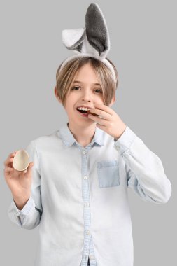 Tavşan kulaklı şirin çocuk gri arka planda çikolata yumurtası yiyor. Paskalya kutlaması
