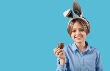 Tavşan kulaklı şirin çocuk mavi arka planda çikolata yumurtası yiyor. Paskalya kutlaması