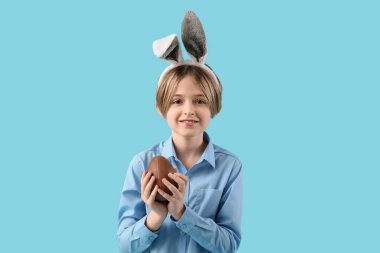 Mavi arka planda çikolatalı yumurtası olan tavşan kulaklı mutlu bir çocuk. Paskalya kutlaması