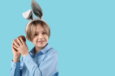 Mavi arka planda çikolatalı yumurtası olan tavşan kulaklı mutlu bir çocuk. Paskalya kutlaması