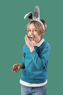 Tavşan kulaklı şirin çocuk yeşil arka planda çikolata yumurtası yiyor. Paskalya kutlaması