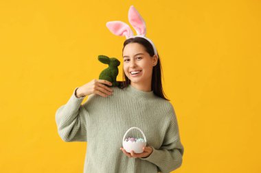 Tavşan kulaklı güzel bir genç kadın, oyuncak tavşan ve sarı arka planda Paskalya sepeti.