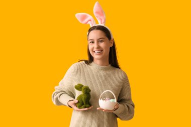 Tavşan kulaklı güzel bir genç kadın, oyuncak tavşan ve sarı arka planda Paskalya sepeti.