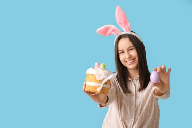 Tavşan kulaklı güzel bir kadın, mavi arka planda Paskalya pastası ve yumurta.