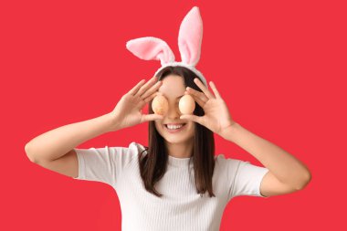 Tavşan kulaklı ve kırmızı arka planda Paskalya yumurtaları olan komik genç bir kadın.