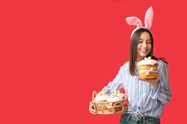 Tavşan kulaklı güzel bir kadın, kırmızı arka planda Paskalya sepeti ve pasta.