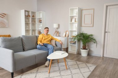 Oturma odasındaki gri koltukta oturan genç adam.