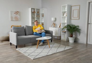 Oturma odasındaki gri koltukta oturan yastıklı genç adam.